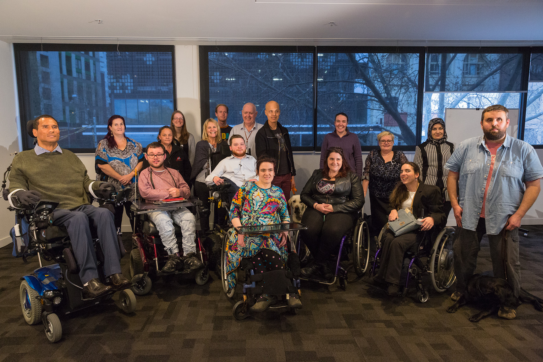 photo of a group of 17 people (and 2 guide dogs) facing the camera, in a conference room. They are in front a a wall of windows - semi transparent blinds are down but a tree and buildings are visible in the background. There is dark grey carpet in the foreground. Our champions team's expressions appear excited and proud.