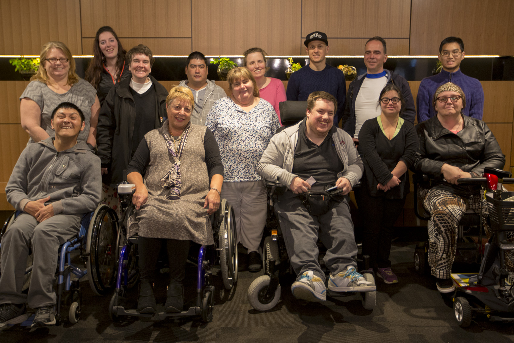 Photo of a group of 14 people in the lunch room at a hotel. They are in front a wood panel wall. There is dark grey carpet in the foreground. The NDIS Champions are smiling and appear relieved after a long day of training.
