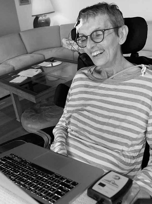 Janet sits in her wheelchair, smiling at the laptop in front of her. She has electrical sensors on her forehead, and a device next to her laptop - these are a part of the NeuroSwitch that allow her to communicate through her computer.
